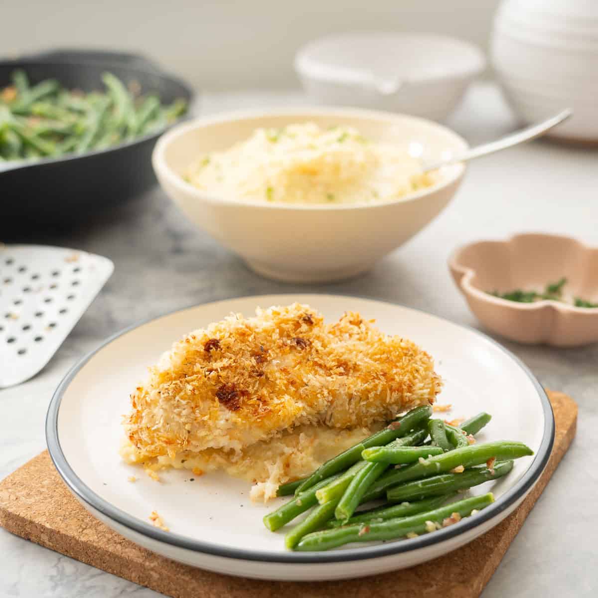 A dinner plate with green beans to the front and a crumbed chicken cutlet resting on mashed potato with a bowl of mashed potato in the background
