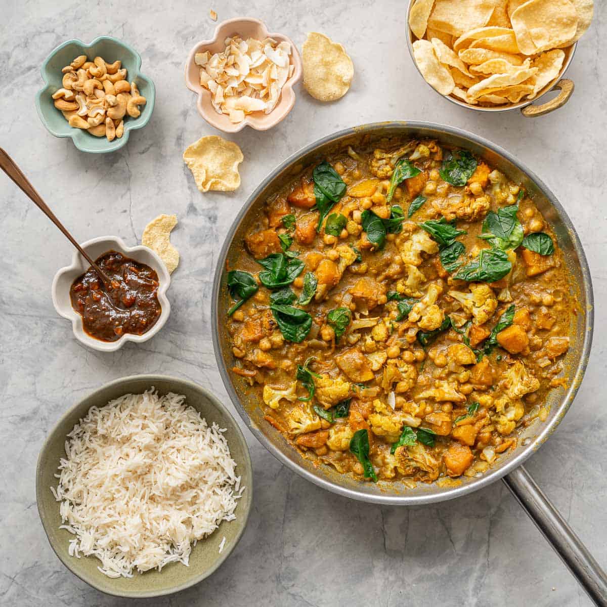 A skillet of chickpea and pumpkin curry next to a bowls of rice, poppadoms, relish, coconut flakes and cashews. 