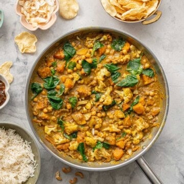 A skillet of chickpea and pumpkin curry next to a bowls of rice, poppadoms, relish, coconut flakes and cashews.