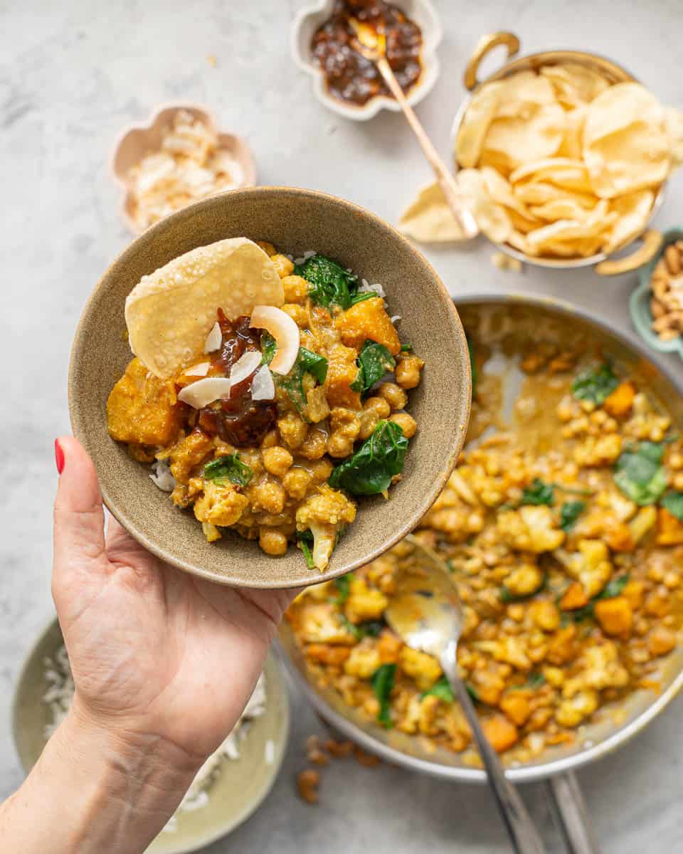 A bowl of chickpea curry garnished with relish and poppadoms being held above a skillet of curry.