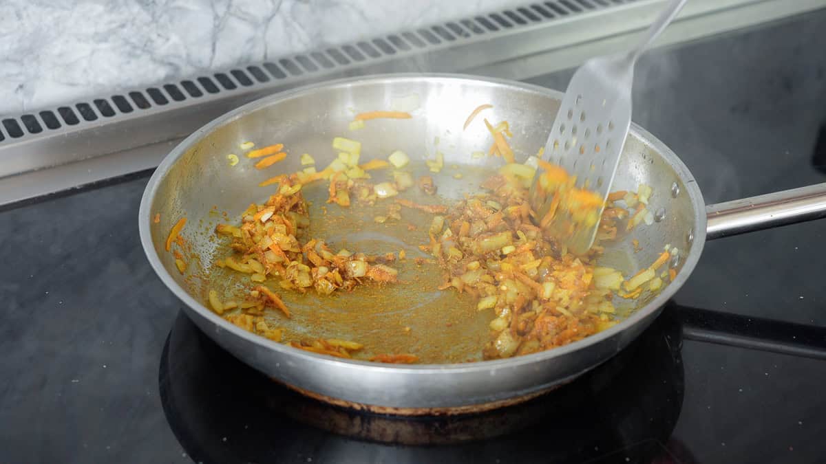 Onion, garlic, grated carrots and spices being pan fried in a stainless steel skillet. 