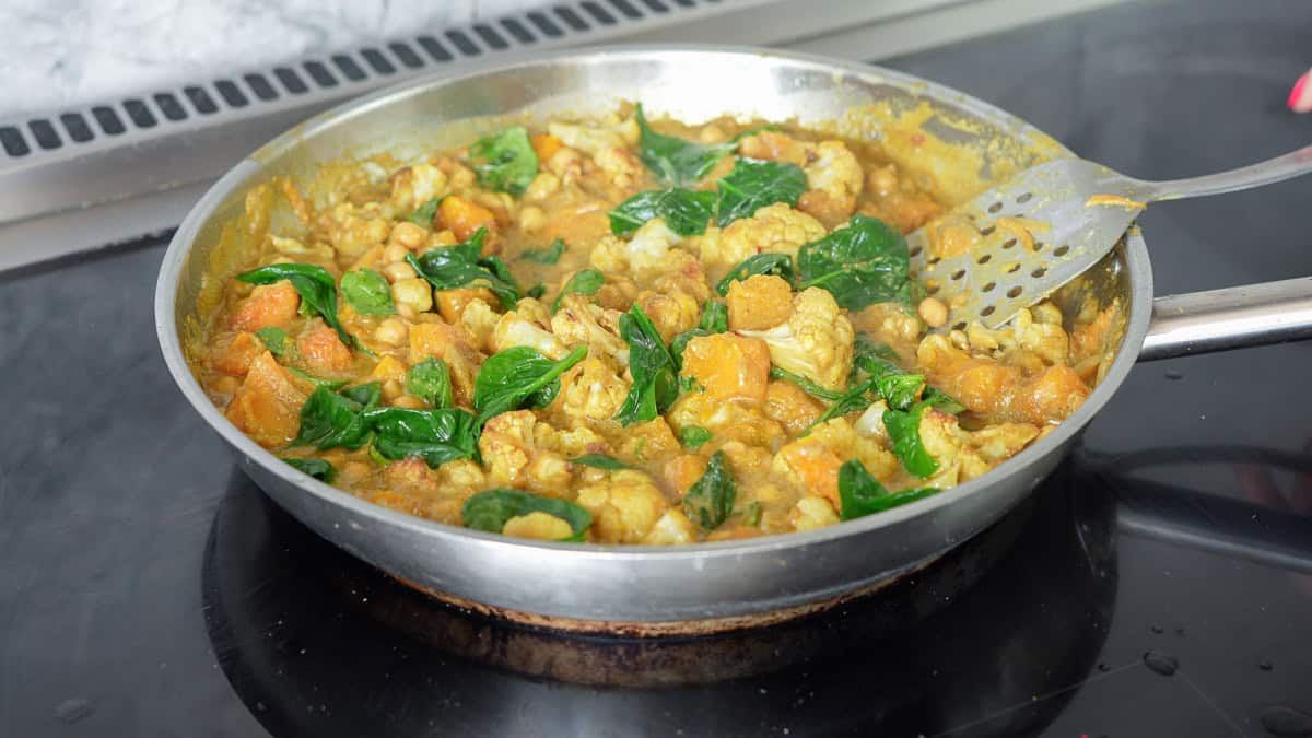 A skillet of curry cooking on a stovetop, chickpeas, cauliflower, spinach and pumpkin pieces visible. 