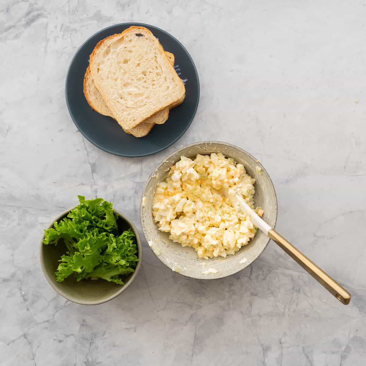 A bowl of egg salad next to a plate of four pieces of bread and a bowl of frilly lettuce leaves. 