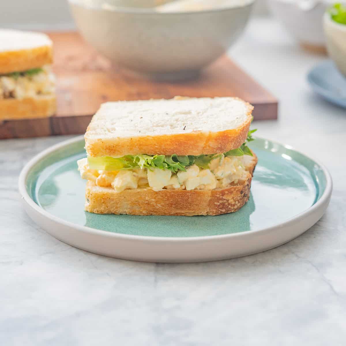 An egg salad and lettuce salad on a turquoise ceramic plate with a second sandwich and a ceramic bowl in the background.