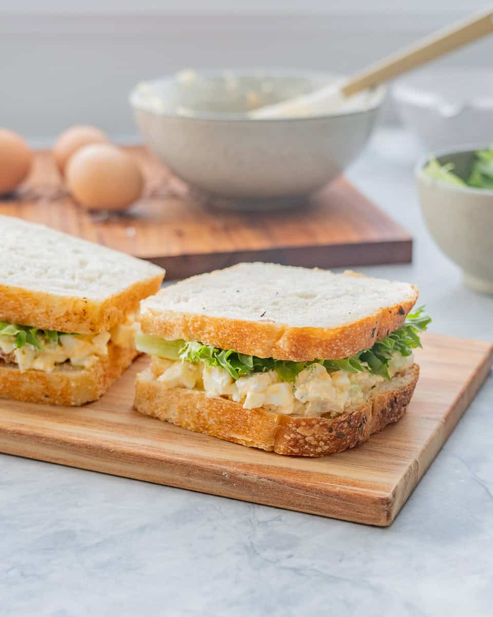 Two egg salad and lettuce sandwiches on a wooden chopping board in front of a bowl of egg salad and boiled eggs. 