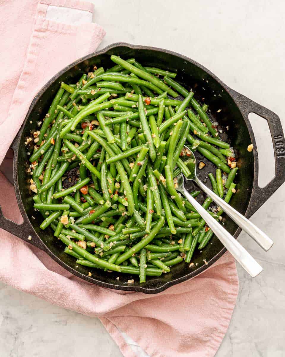 A cast iron skillet of garlic butter coated green beans draped with a pink tea towel sitting on a grey marble bench top. 