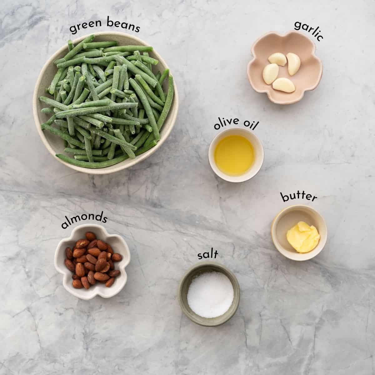 The ingredients to make garlic green beans laid our on a bench top in bowls with text overlay ingredient name labels. 
