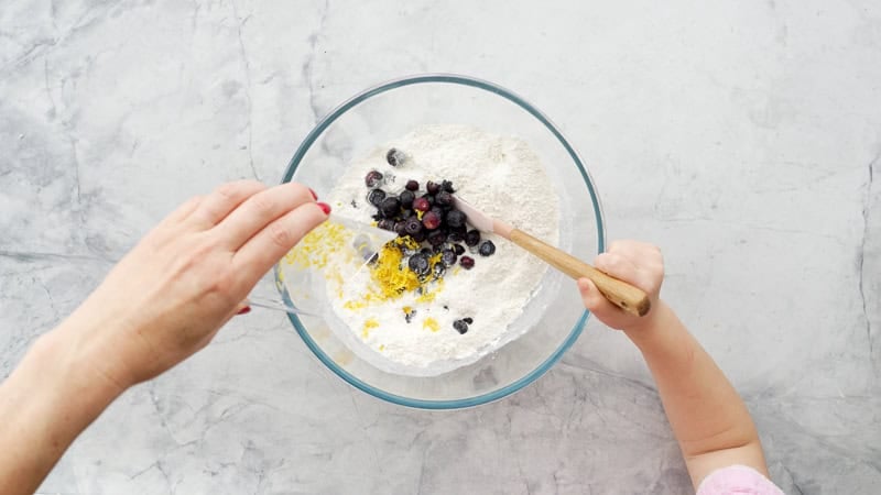 Self-raising flour, blueberries, and lemon zest placed in a glass bowl on a bench and being mixed with a wooden spoon