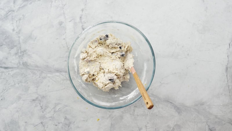 The scone dough mixed together in a glass bowl resting on the bench with a spatula resting against the side. 