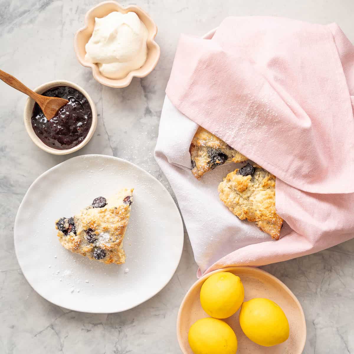 One baked blueberry scone resting on a plate next to the remainder of the batch of scones which is resting in a soft pink tea towel and one ramekin filled with jam and another with whipped cream and a plate with three lemons 