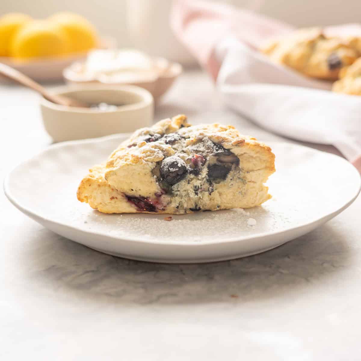 A triangular shaped blueberry scone on a side plate with bowls of cream, sauce and lemons in the background.
