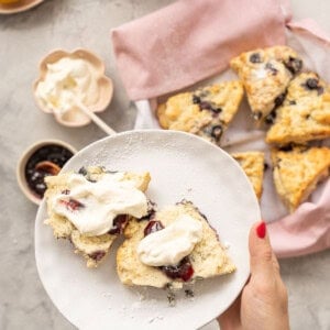 A blueberry scone topped with cream above a plate of more scones wrapped in a pink tea towel.