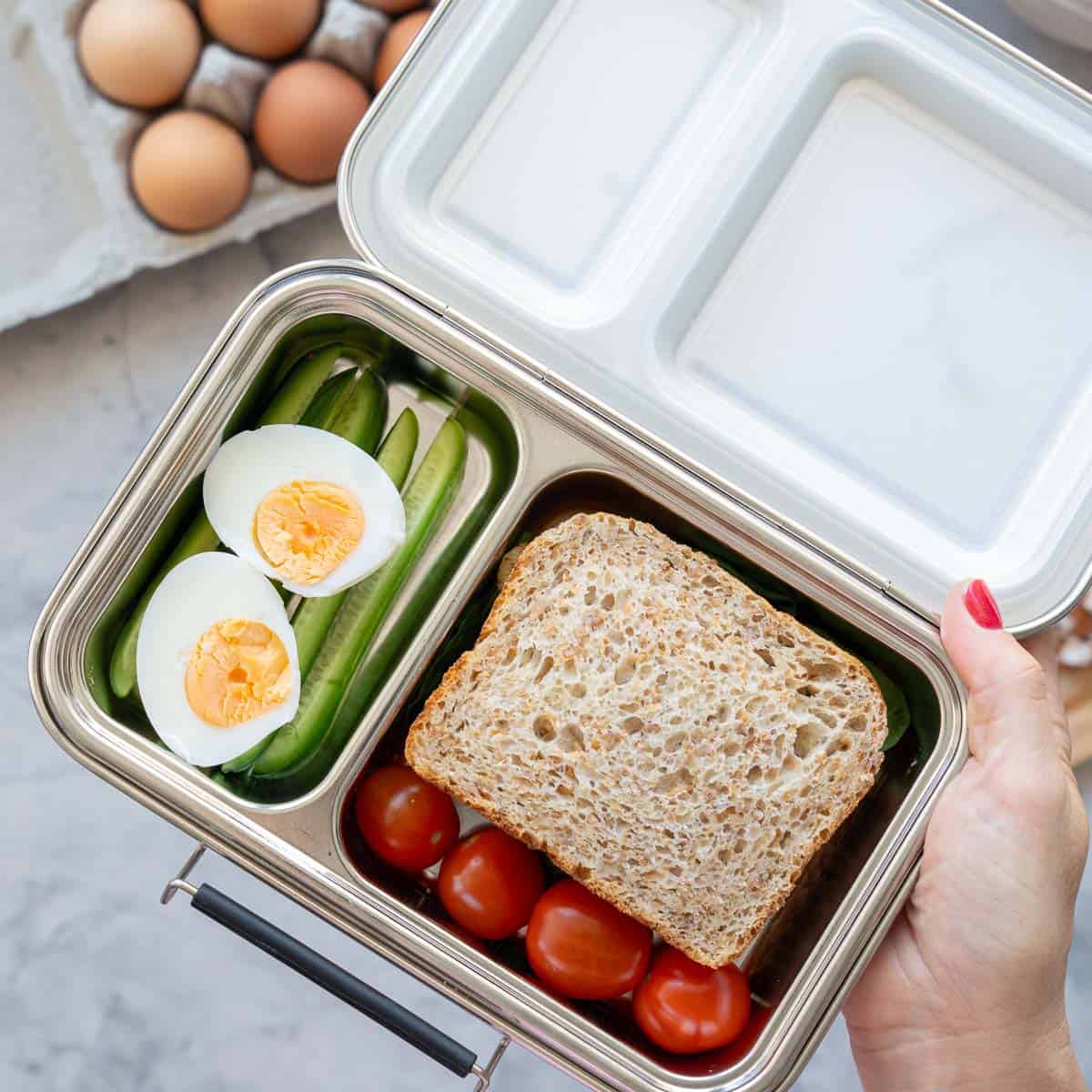 A stainless steel lunch box packed with a sandwich, cucumber sticks and a hard boiled egg cut in half.