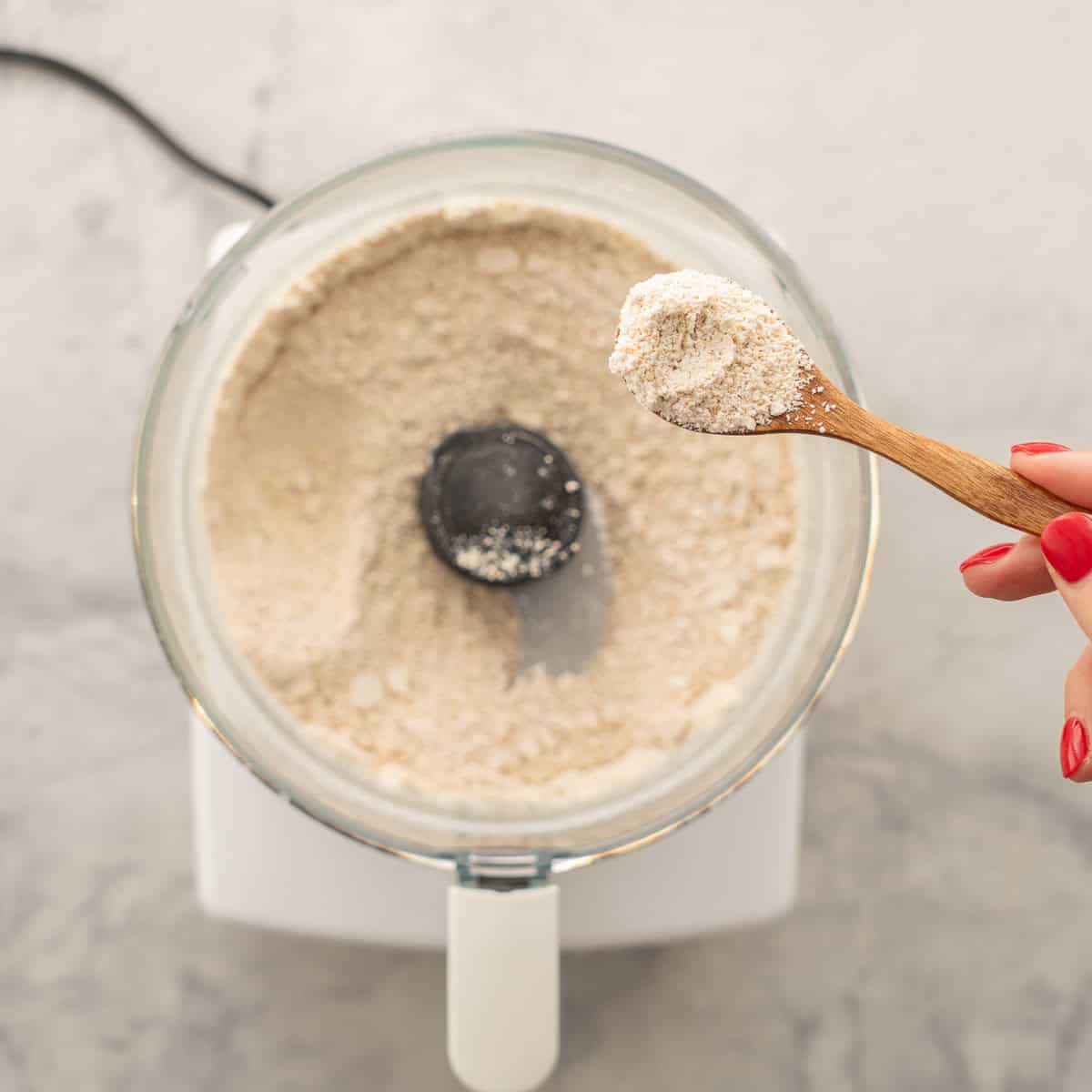 A food processor full of blitzed oats resting on the bench with a hand holding a small wooden spoon with a scoop of the fine oat flour