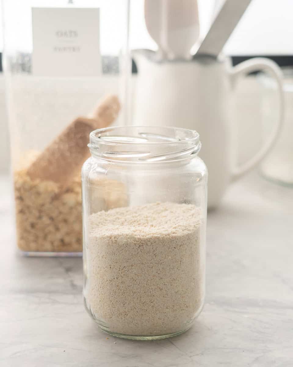 One jar of oat flour resting on the bench in front of a container of oats 