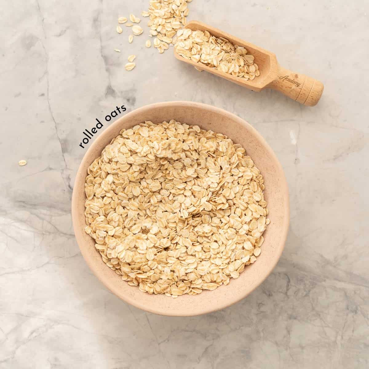 A soft pink ceramic bowl full of rolled oats resting on the bench next to a small wooden scoop of oats