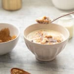 A spoonful of creamy oatmeal being held above a ceramic bowl of oatmeal which is topped with meting brown sugar and cream.