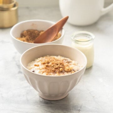 A cream ceramic bowl filled with oatmeal in front of a bowl of brown sugar cinnamon topping and a small glass bottle of cream.