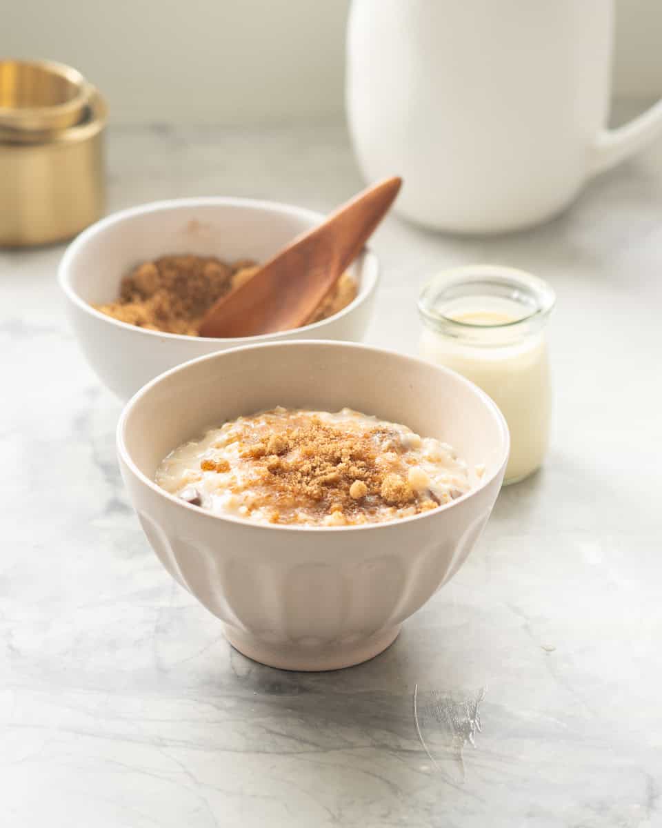 A cream ceramic bowl filled with oatmeal in front of a bowl of brown sugar cinnamon topping and a small glass bottle of cream. 