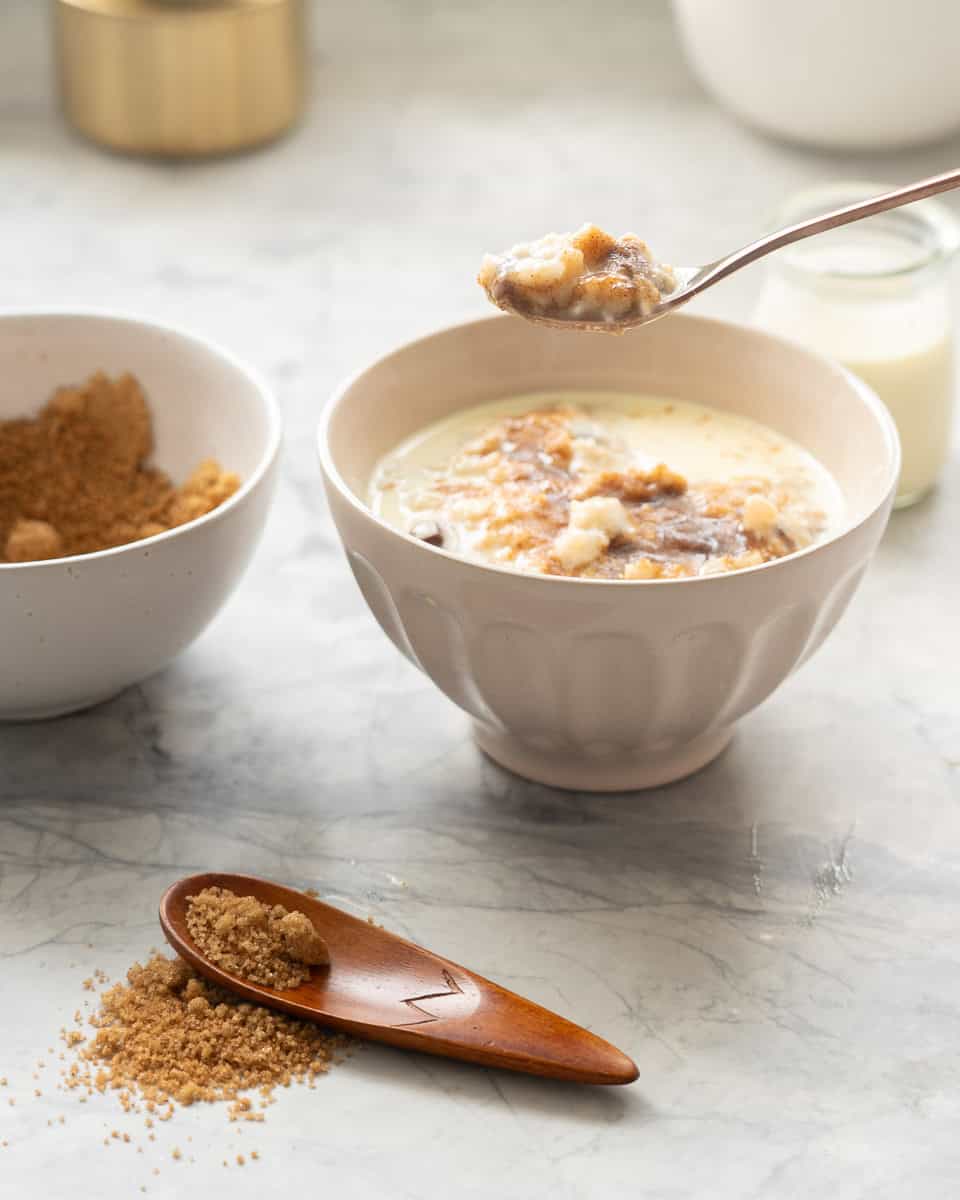 A spoonful of creamy oatmeal being held above a ceramic bowl of oatmeal which is topped with meting brown sugar and cream. 