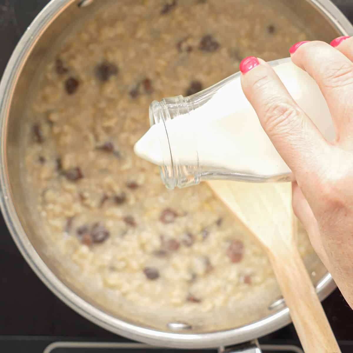 A small glass bottle of milk being poured into a saucepan of cooked oatmeal. 