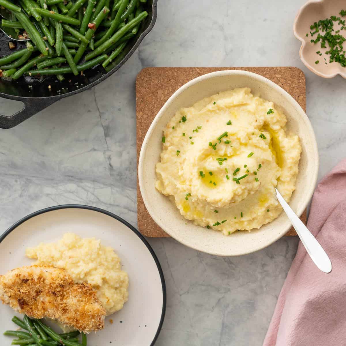 A bowl of potato and cauliflower mash, drizzled with olive oil and sprinkled with chopped chives. next to a dinner plate of crumbed chicken mash and green beans. 