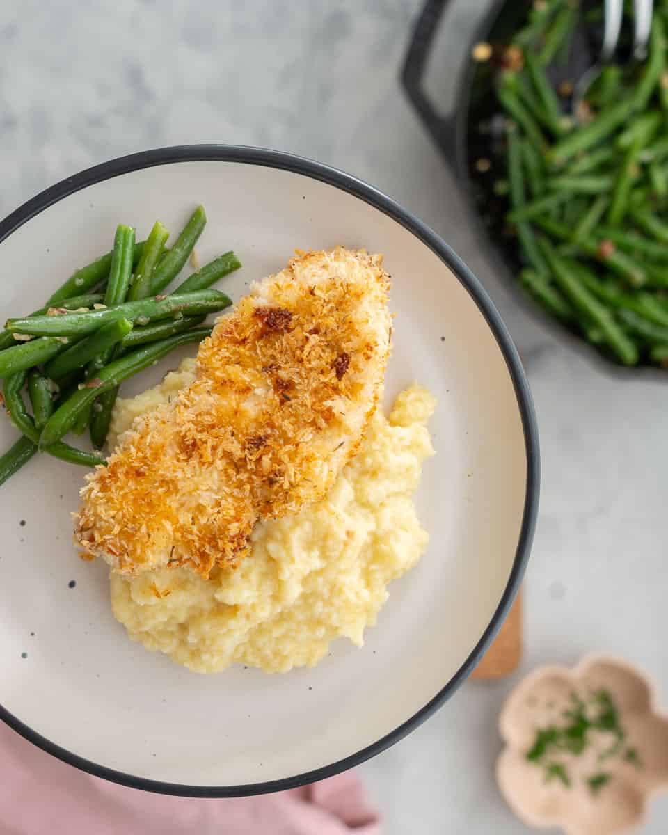 A crumbed chicken cutlet resting on mashed potatoes next to a pile of green beans on a dinner plate. 