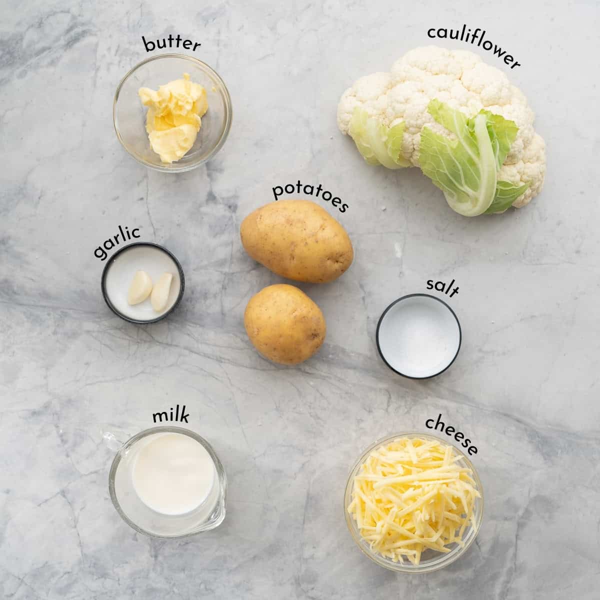 The ingredients to make potato cauliflower mash laid out on a marble bench top with text overlay ingredient labels. 