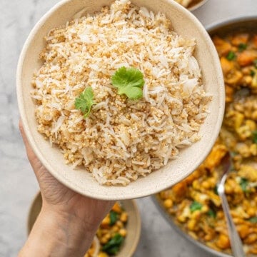 A bowl of cooked rice and quinoa garnished with coriander being held above a skillet of curry.