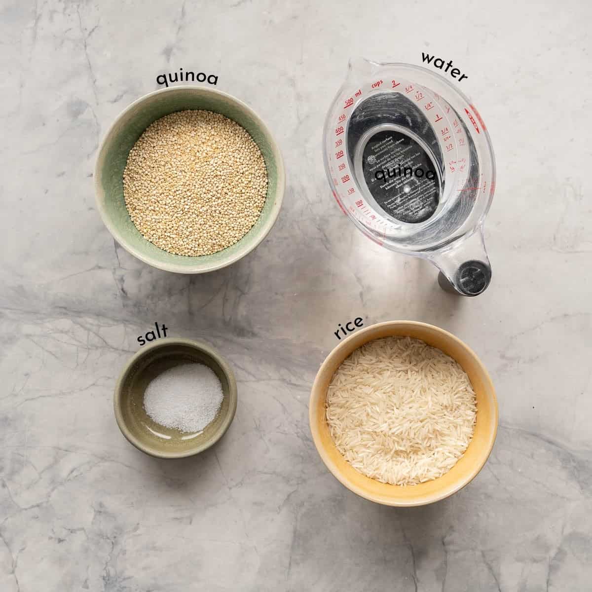 Bowls of quinoa, water, rice and salt each labeled with a text overlay laid out on a bench top. 