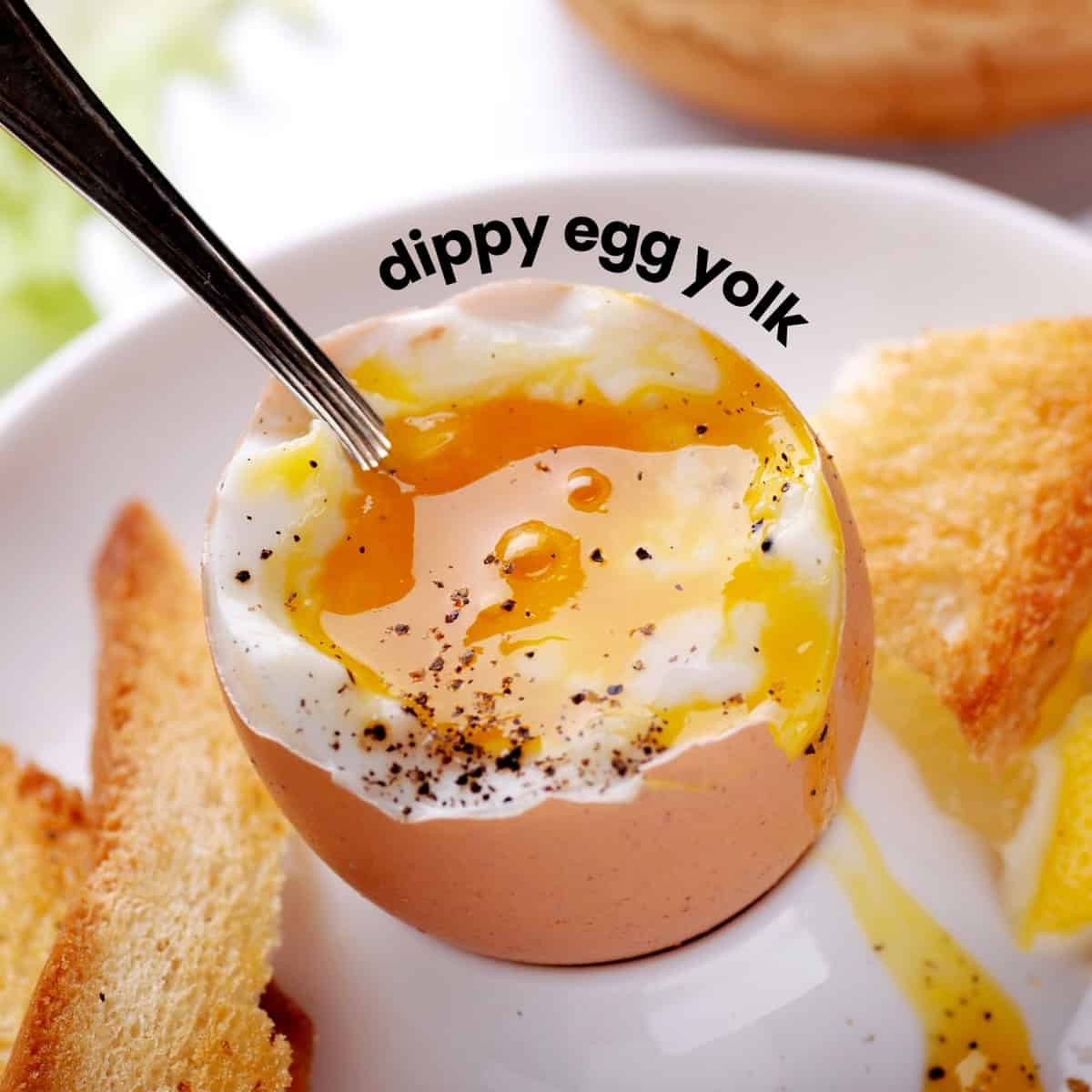 An egg in an egg cup with a spoon resting in its runny egg yolk. Text overlay: dippy egg yolk. 