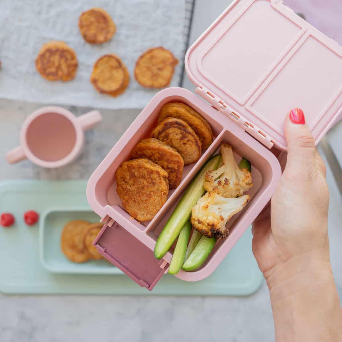 A small pink bento box packed with cauliflower florets, cucumber sticks and mini pancakes.