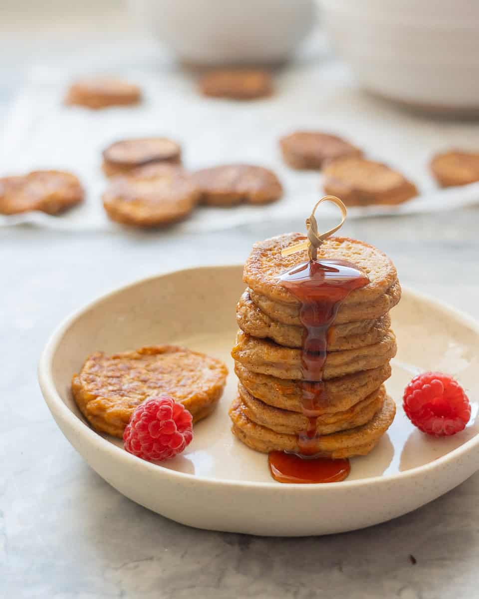 A stack of seven pancakes drizzled with maple on a side plate with two raspberries.