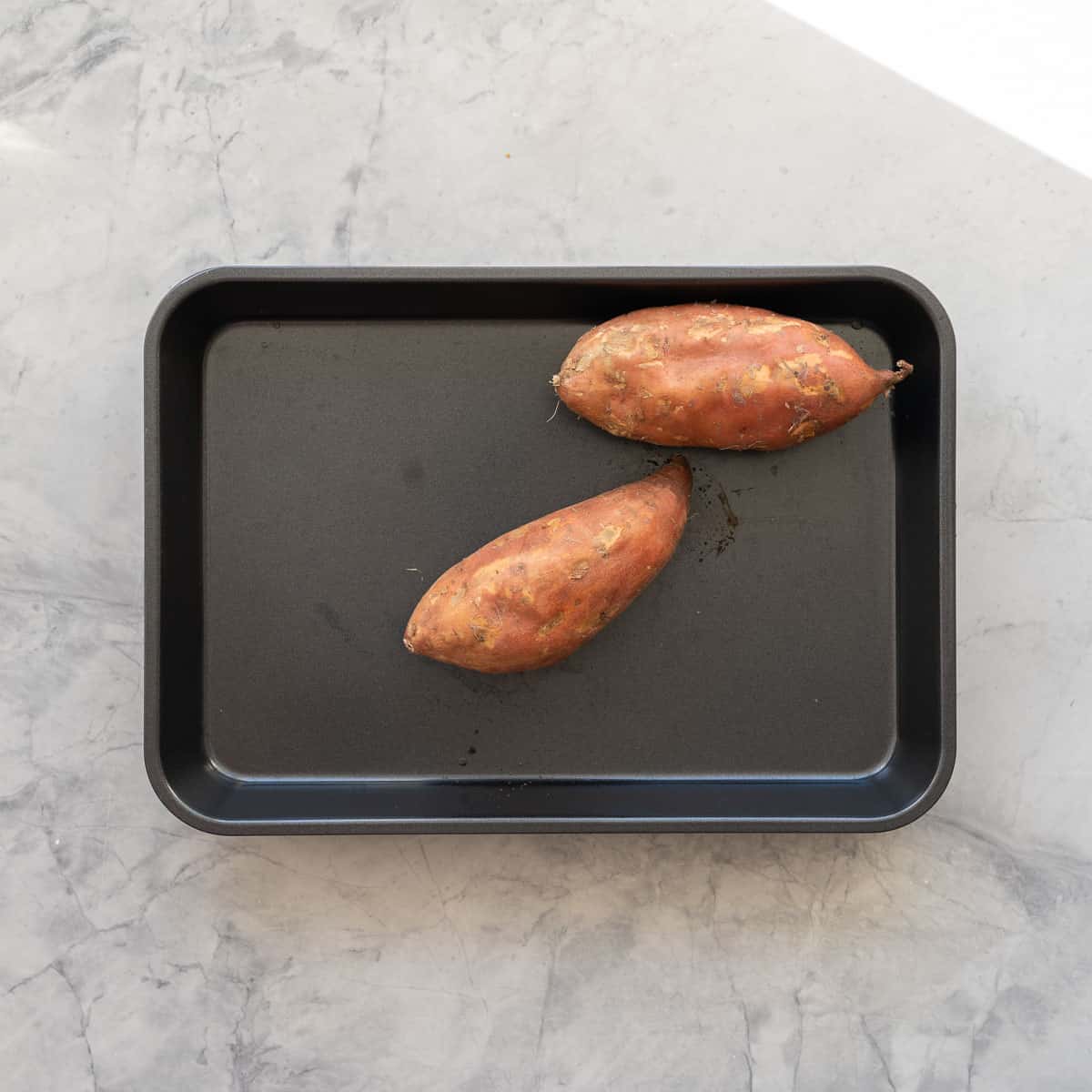Two raw whole sweet potatoes on a baking tray.
