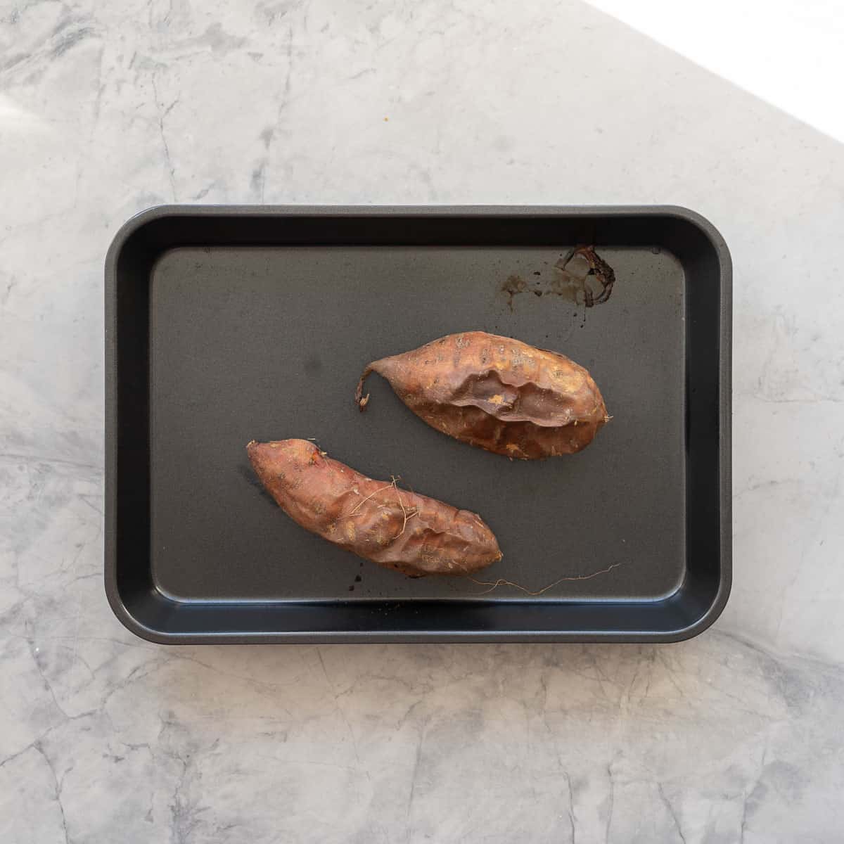 Two roasted sweet potato with their skins on sitting in a on a baking tray.