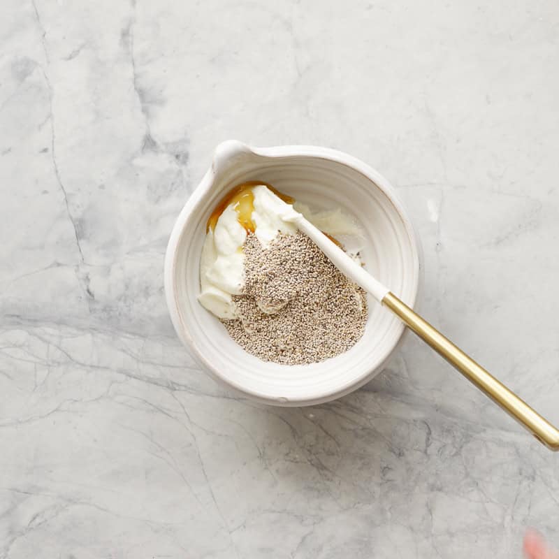 A white ceramic bowl of yogurt, chia seeds and maple syrup ready to be mixed with a spatula. 