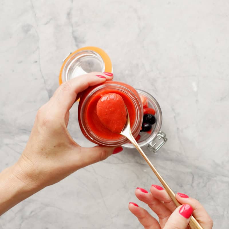 A golden spoon lifting a spoonful of bright red fruit puree from a glass jar into a second jar part filled with berries. 