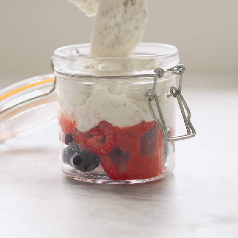 Chia seeds mixed into yogurt being spooned into a glass jar over a layer of mixed berries. 