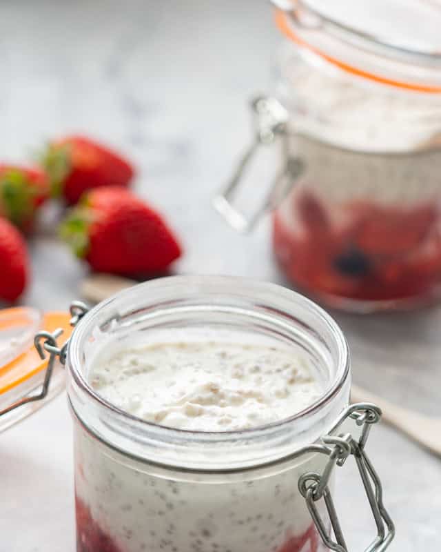 Plump chia seeds in yogurt in a glass jar, strawberries and a chia seed pudding visible in the background. 