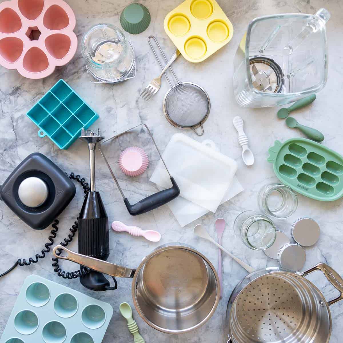 All the cooking utensils associated with making homemade baby food laid out on a bench top, pots, steamers, blender, sieve, ice cube trays, zip lock bags.