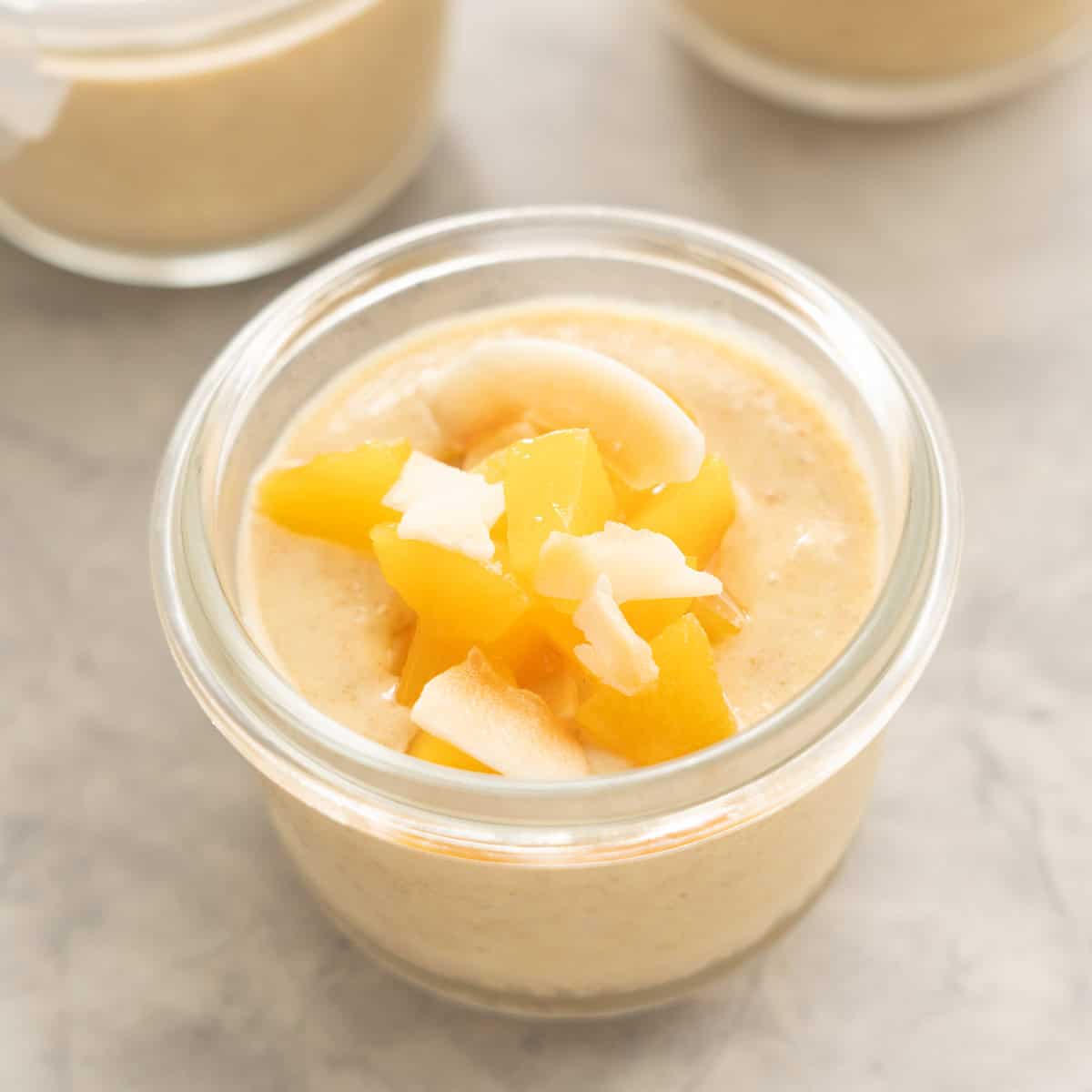 A peach coloured pudding in a glass jar topped with glistening cubes of peach and toasted coconut chips. 