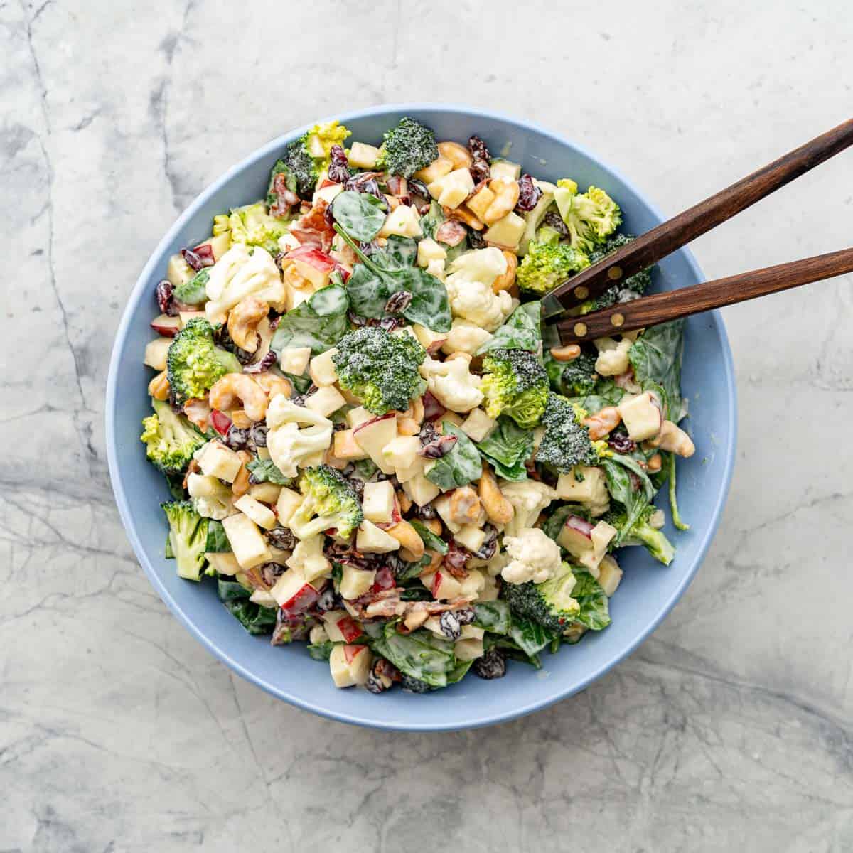 A big blue salad bowl filled with apple spinach broccoli cauliflower raisins and cashews, topped with creamy salad dressing, with wood tongs in bowl.