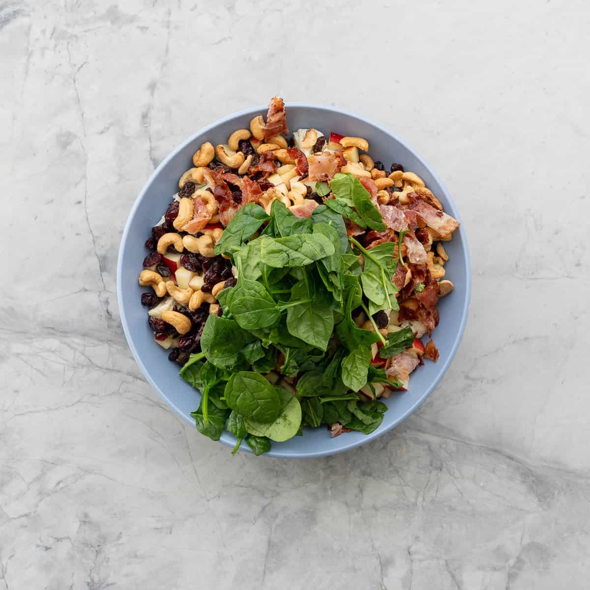 A big blue salad bowl filled with cashews spinach bacon and raisins, not mixed.