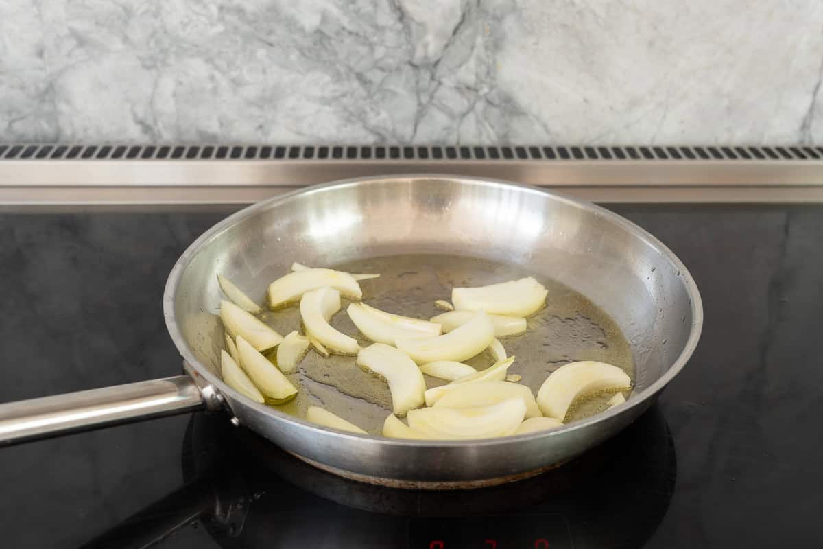 A large fry pan on the cooktop sauteing onions in oil.