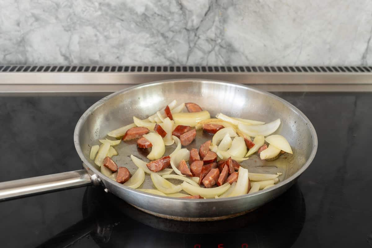 A large fry pan on the cooktop sauteing onions  and chorizo in oil.
