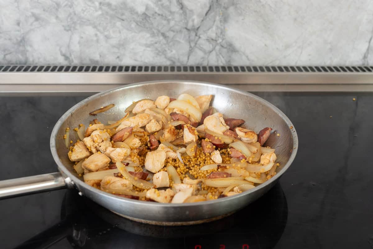 A large fry pan on the cooktop with onions, chorizo, chicken and  couscous simmering.