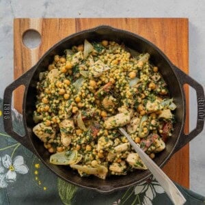 A large cast iron skillet of Chicken Couscous resting on a wooden chopping board on a bench.