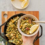 A large cast iron skillet of Chicken Couscous resting on a wooden chopping board on a bench with a hand holding a serving on a plate above it.