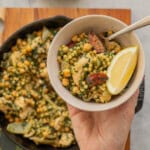 A large cast iron skillet of Chicken Couscous resting on a wooden chopping board on a bench with a hand holding a serving on a plate above it.
