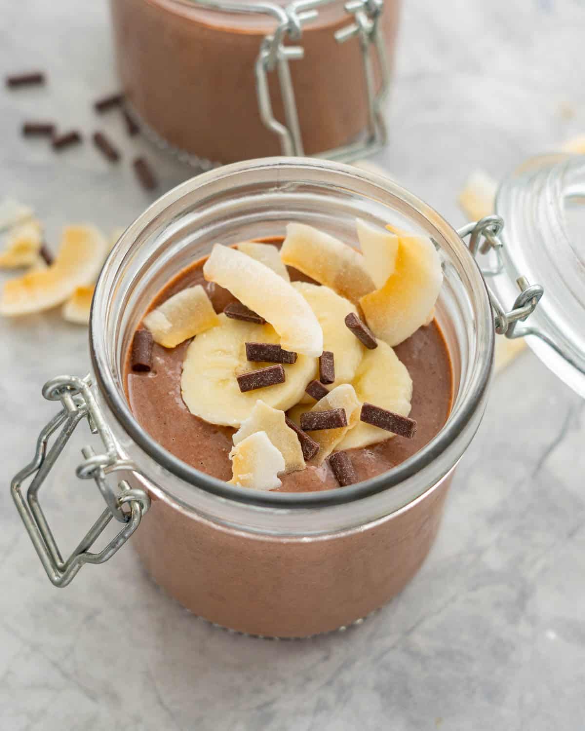A chocolate pudding in a jar topped with banana slices, coconut chips and chocolate chips. 